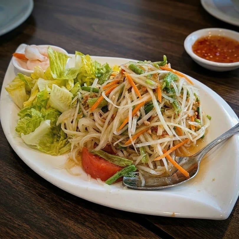 A vibrant Thai papaya salad with green beans, carrots, and lettuce, served with sauce.